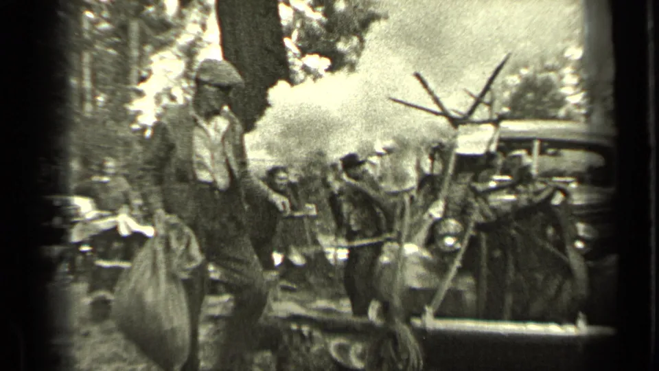 A man in cap and work clothes carries a large sack in the Bend, Oregon camp scene, with smoke, tubs, and other adults behind him.