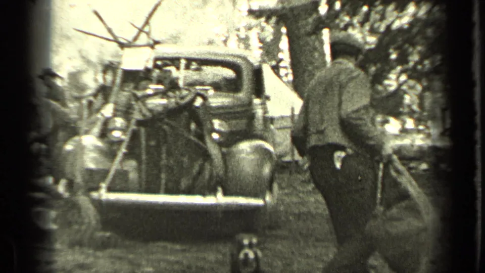In the supplied 1937 Bend, Oregon footage, a man in work clothes carries a large bag near a vintage truck, with a dog and tent visible.
