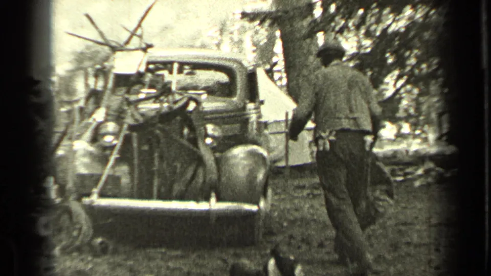 A man walks through a wooded Bend, Oregon campsite near a 1930s-style truck, with a canvas tent and camp equipment in the background.