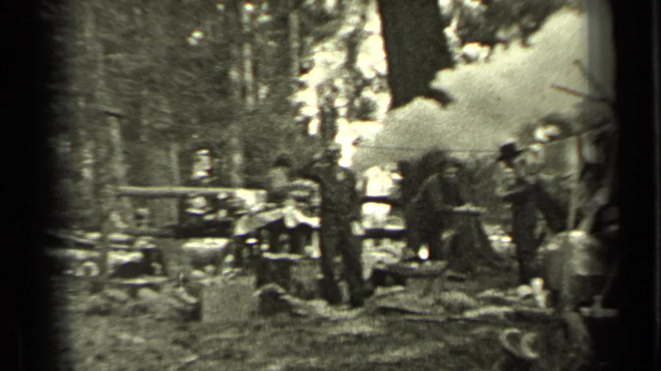 A wider view shows several men at a wooded Bend, Oregon camp, with smoke rising near the work area and a vintage truck partly visible.