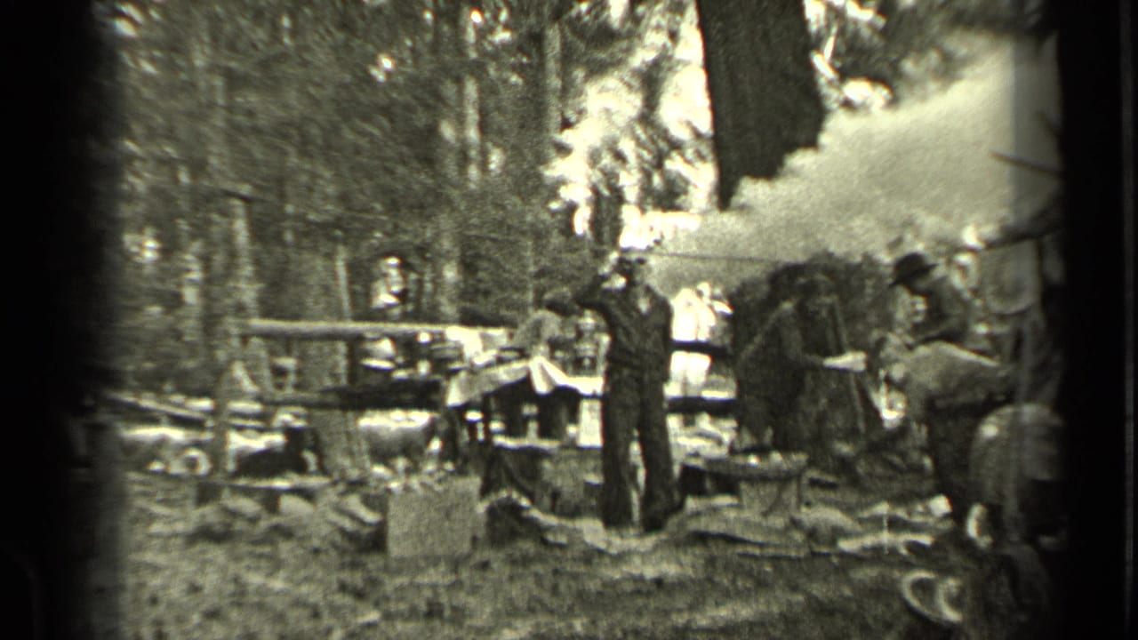 People gathered in a wooded campsite near smoke, a picnic table, and scattered wood.