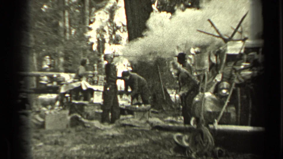 1937 Bend, Oregon campsite frame with a man near a smoky area, other adults nearby, and trees surrounding the scene.
