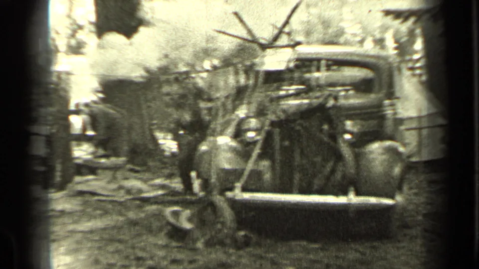 1937 Bend, Oregon home-movie frame showing a vintage car at a wooded campsite with smoke and people in the background.