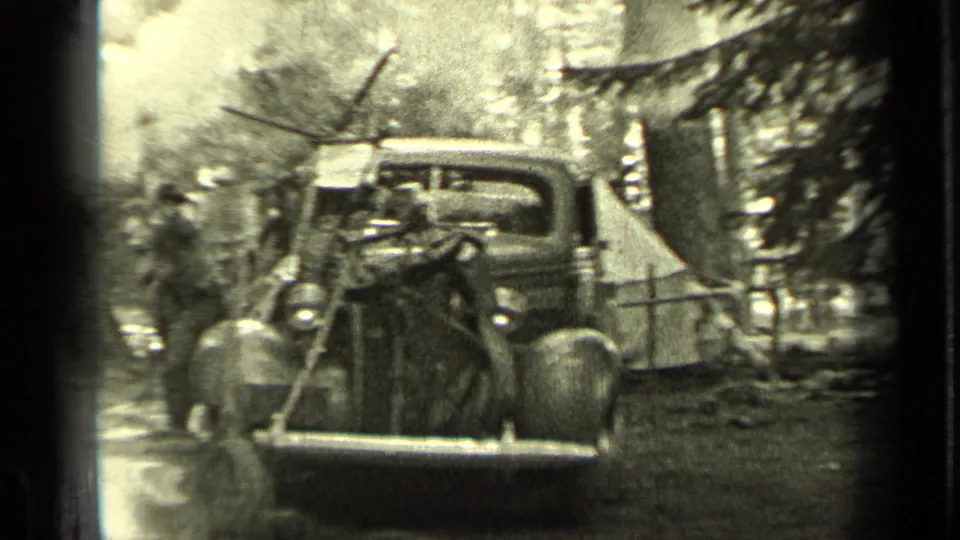 1937 Bend, Oregon frame with a front view of a parked vintage car, adults nearby, smoke, trees, and rough campsite ground.