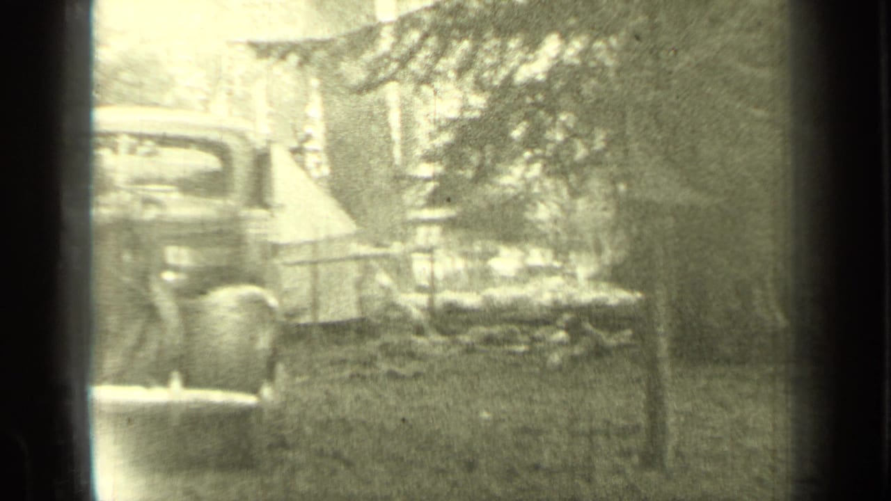Wooded campsite with a tent, parked older automobile, and trees seen through film gate edges.
