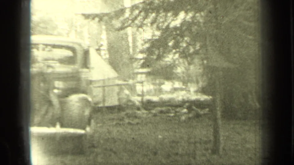 1937 Bend, Oregon home-movie frame showing a tent, older automobile, trees, and brush at a wooded campsite.