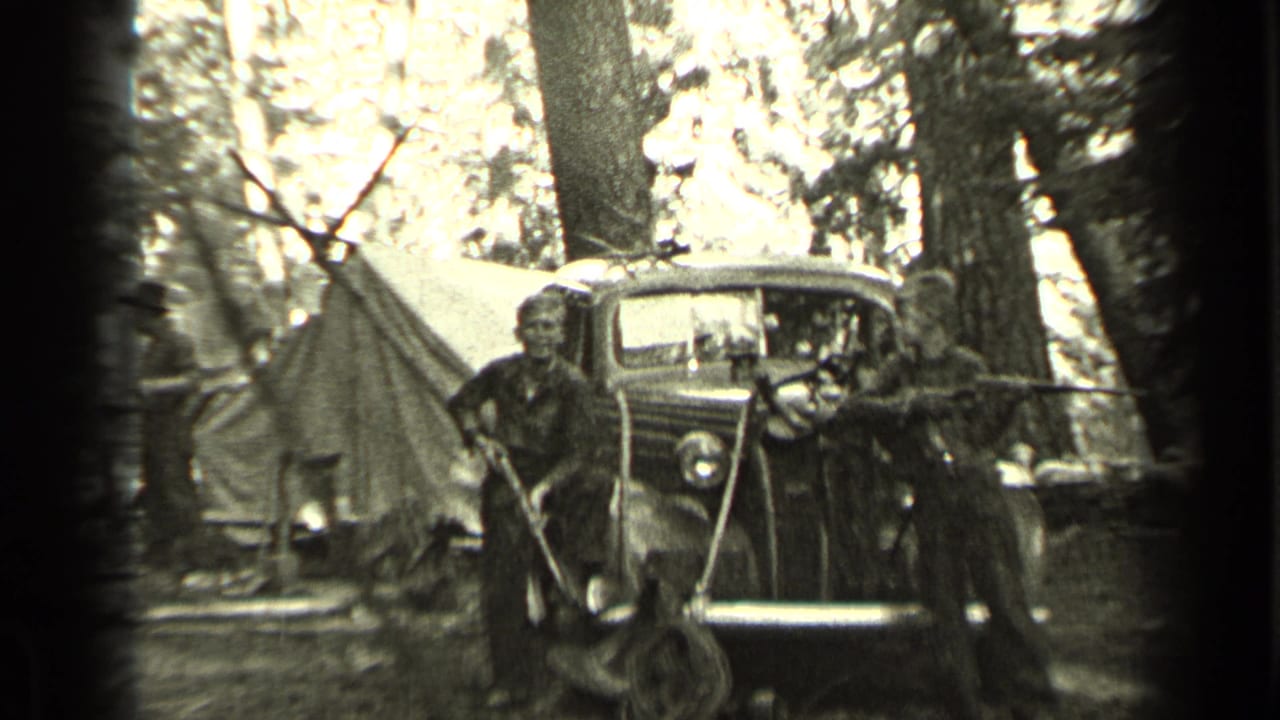 Two boys holding rifles stand by a vintage car and tent in a wooded campsite.