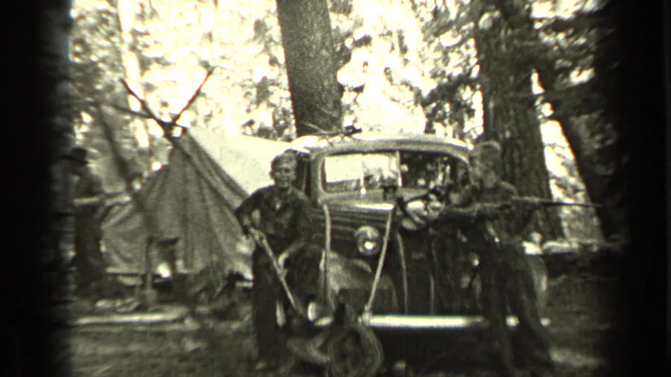 Two boys with rifles pose near a vintage car and canvas tent at a wooded Bend, Oregon campsite in 1937 home-movie footage.