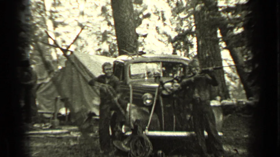 A boy holding a rifle stands near a canvas tent, vintage car, and dogs in 1937 Bend, Oregon campsite footage.