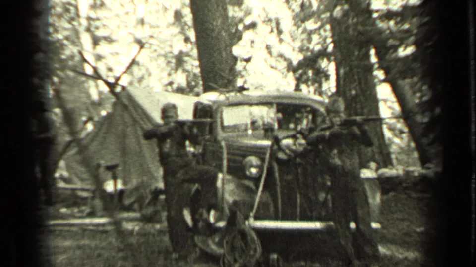 A boy with a rifle stands before a vintage car as dogs gather nearby at the wooded Bend, Oregon campsite in 1937 footage.