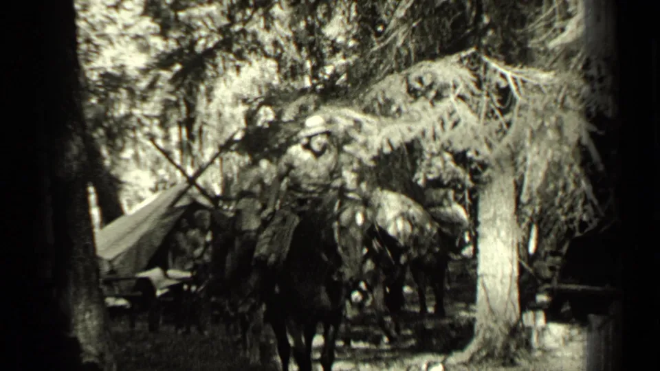 In this 1937 Bend, Oregon home-movie frame, mounted men and loaded horses pass through a forested area with canvas camp gear nearby.