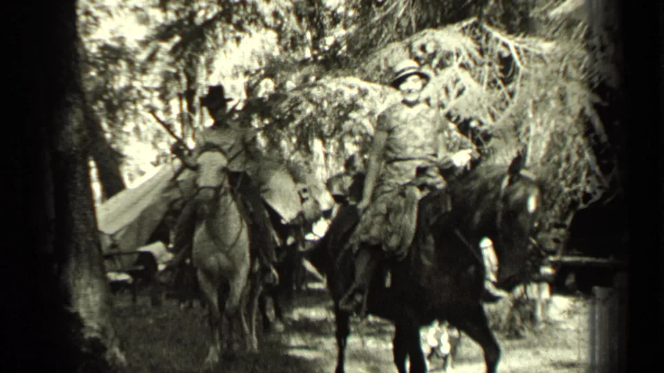 A group of riders on horses, some with visible hats, moves through a wooded Bend, Oregon setting with camp equipment in view.