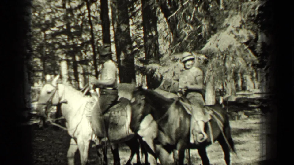 In the Bend, Oregon home-movie scene, two adult riders sit on a light horse and a darker horse beneath tall trees.