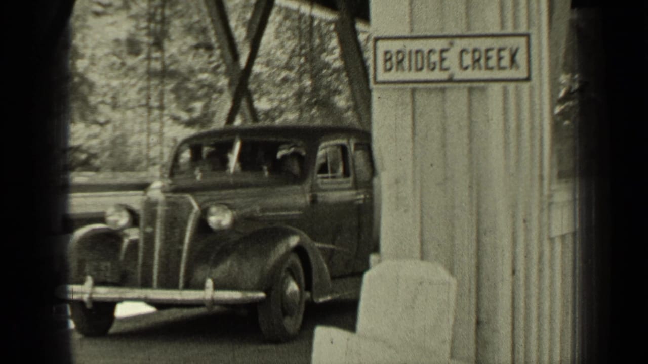 Period car passing through a covered bridge beside a Bridge Creek sign.