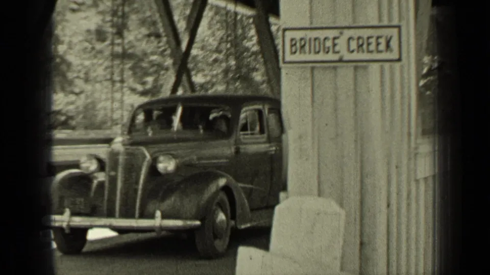 1937 Oregon home-movie frame showing a period automobile at a covered bridge with a visible Bridge Creek sign.