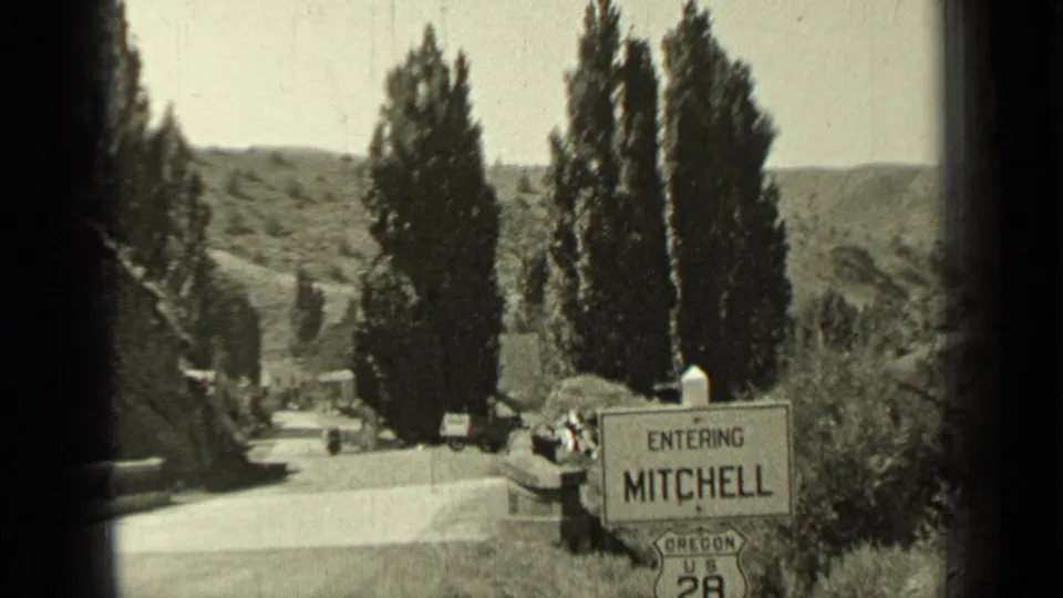 1937 Oregon travel frame showing the Entering Mitchell sign on US 28, with cars, trees, hills, and possible distant people nearby.