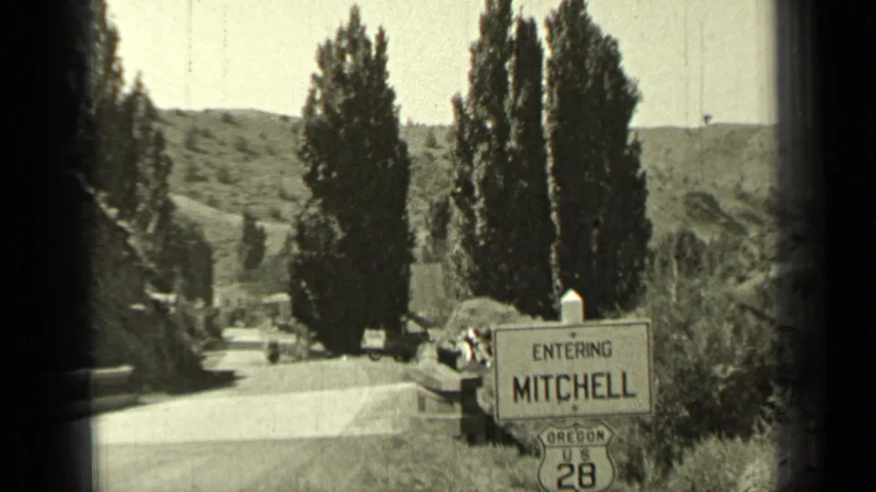1937 Oregon home-movie frame showing an Entering Mitchell road sign near tall trees, a quiet road, and parked cars.