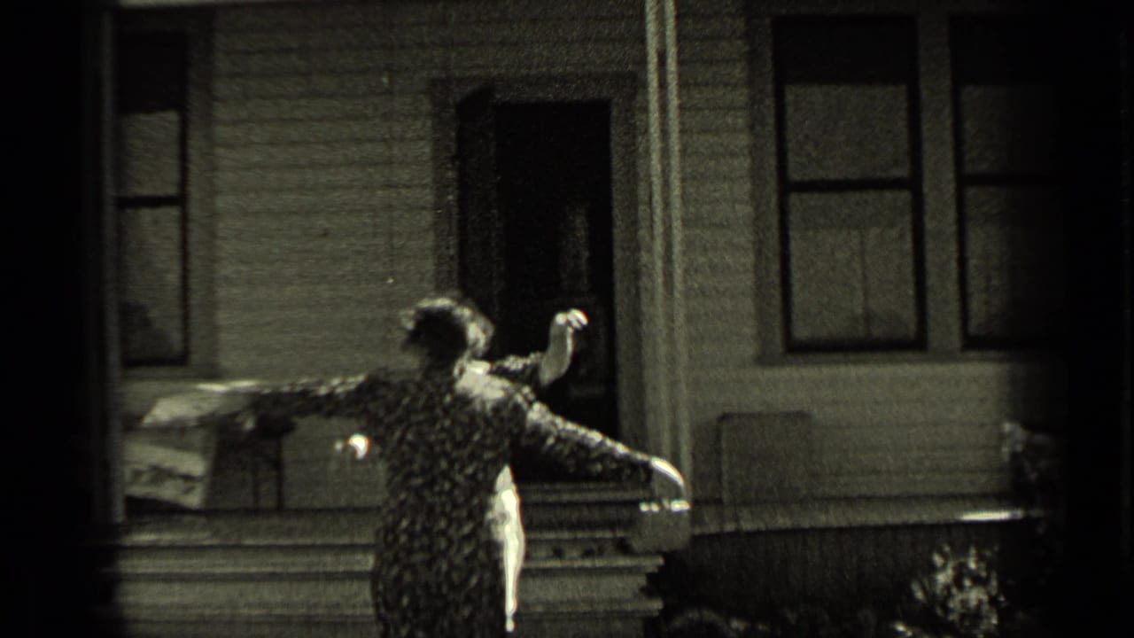 A woman in a patterned dress stands outside a wood-sided house in Oregon, opening her arms in greeting.