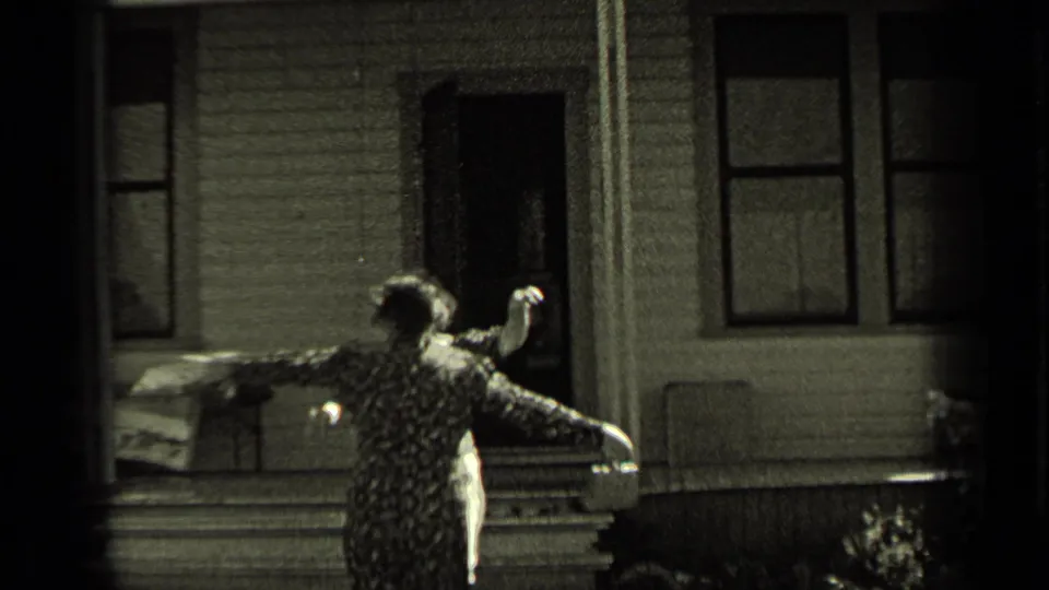A woman in a patterned dress stands outside a wood-sided house in Oregon, opening her arms in greeting.