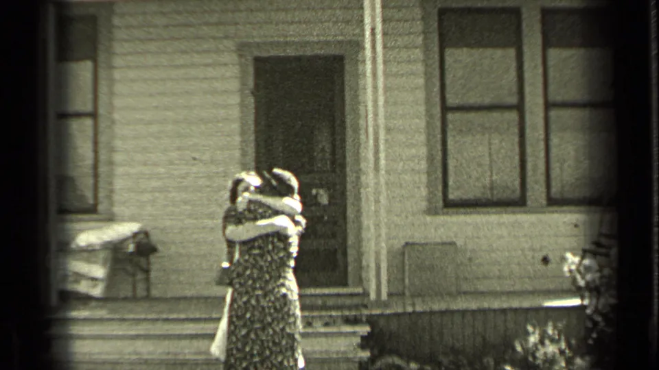 Two women embrace outside a modest wood-sided house, continuing the home-movie reunion scene in Oregon.