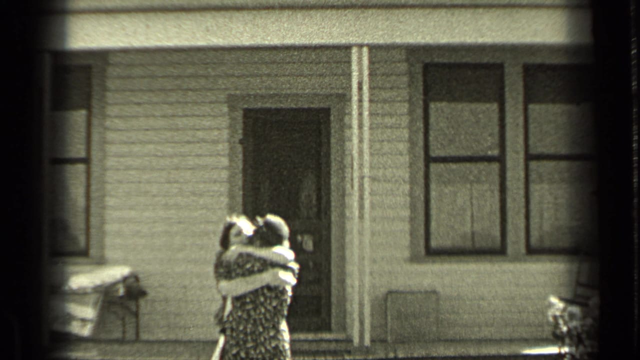 Two people embrace or stand close outside a house with porch and windows.