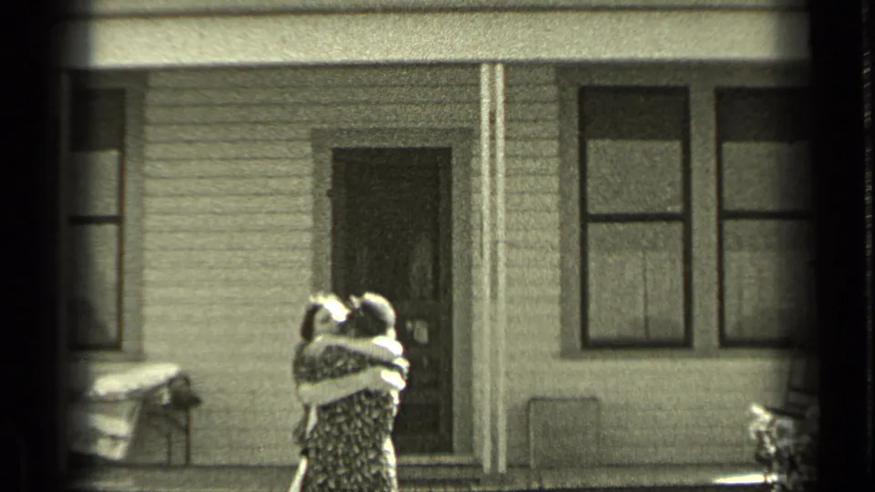 In the supplied 1937 Bend, Oregon home-movie context, two people stand close together outside a house facade with porch details and windows.