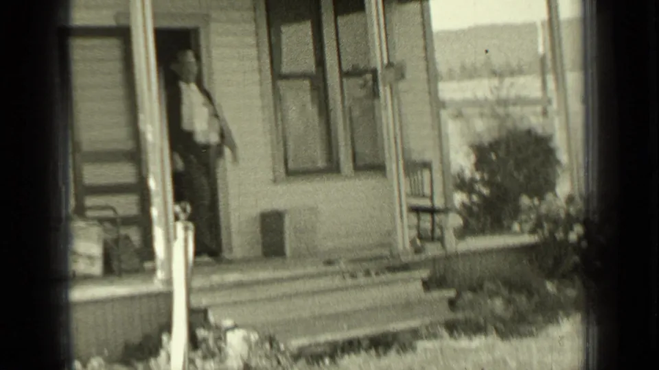 A person stands on the porch of a weathered house in the 1937 Bend, Oregon ranch scene, with windows, a chair, and open land nearby.