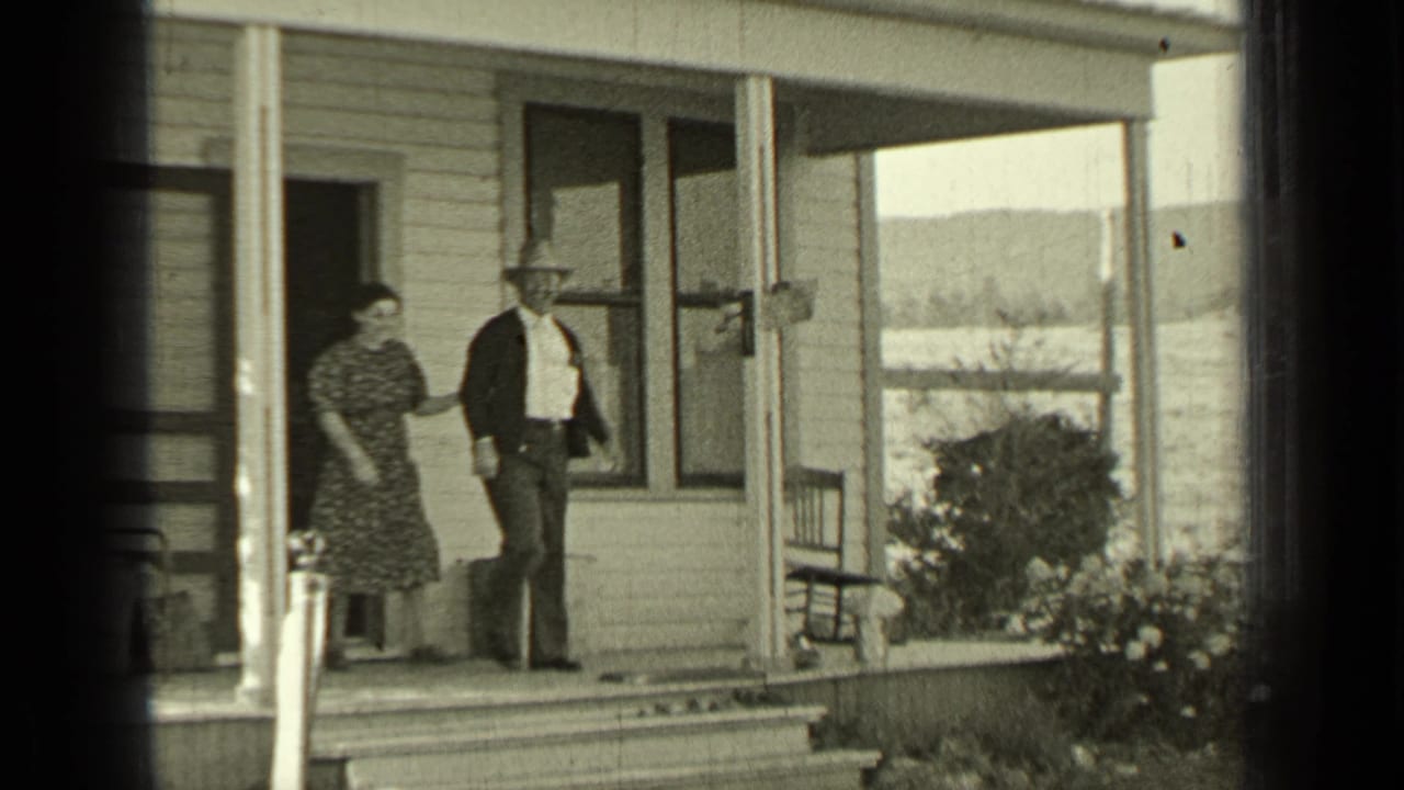 Man and woman standing on a farmhouse porch with fields beyond.