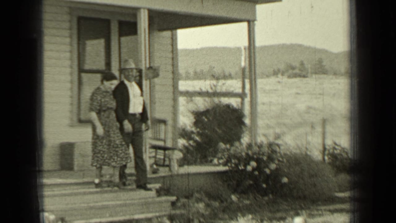 Man and woman stepping from the porch beside bushes and open fields.