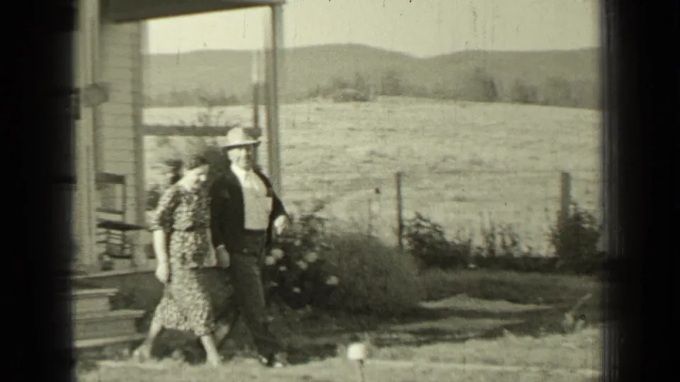 A man and woman walk closely beside the farmhouse, with porch steps, flowering bushes, open pasture, and low hills visible in the background.