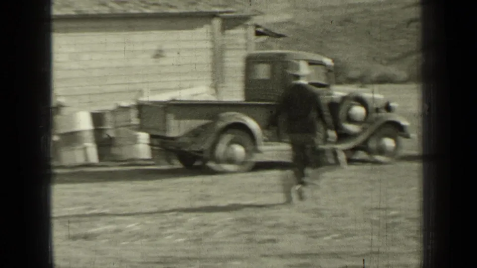 Motion blur shows the man crossing the yard toward the pickup, with the wooden building and open landscape behind him.