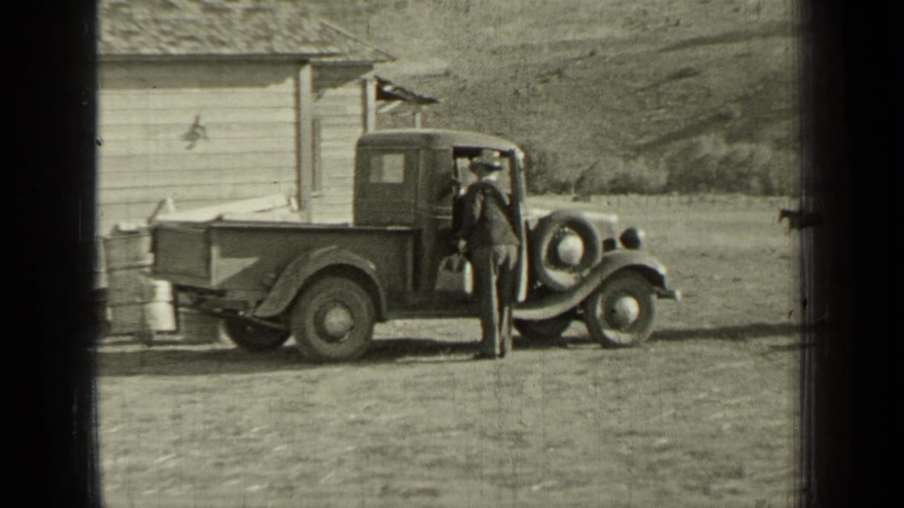 Man in a suit stands beside a vintage pickup near a wooden building.