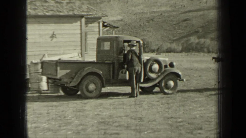 In Bend, Oregon, 1937, a suited man stands by the open door of a vintage pickup in a rural yard.