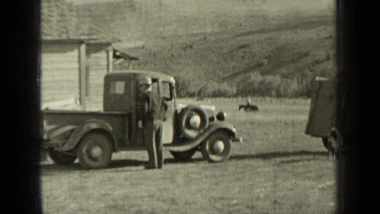 Man stands at the driver side of a vintage pickup in a rural yard.