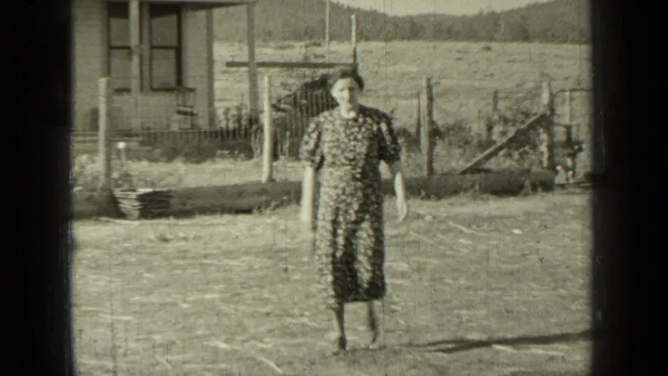 A woman in a patterned dress walks toward the camera in a rural Bend, Oregon yard, with fencing and a building behind her.