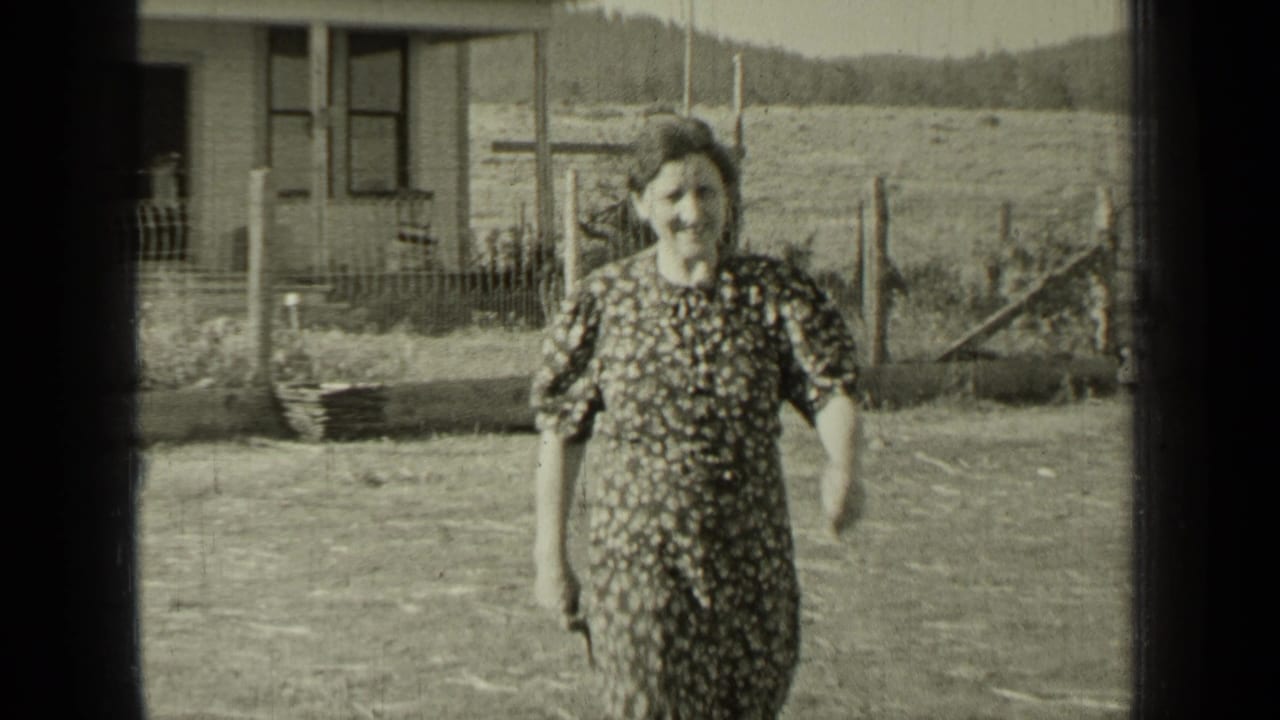Woman in a patterned dress walks across a rural yard near a house and fence.