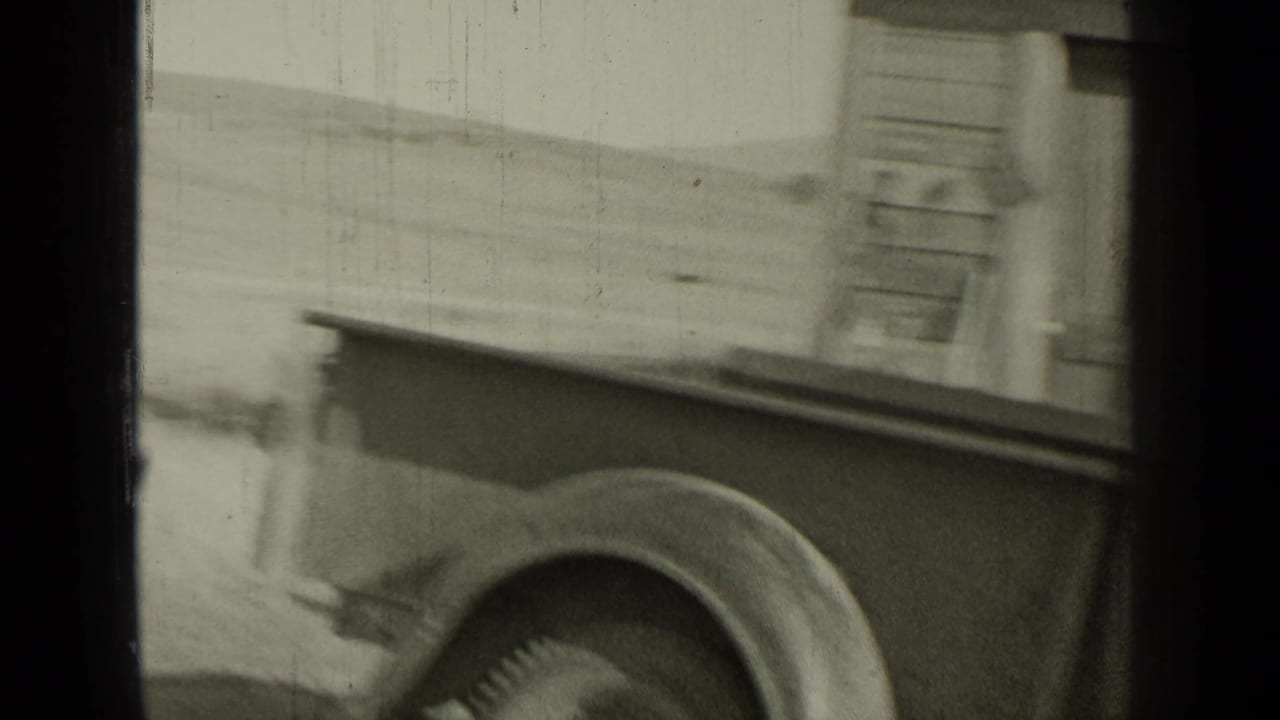 Side of a vintage vehicle beside a building, with the rear wheel and body visible.