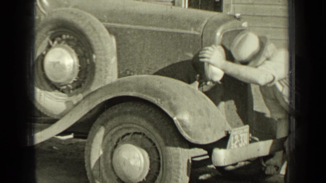 Man in a hat cranking the front of a vintage automobile beside rounded fenders and spoked wheels.