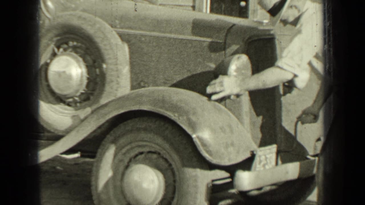 Man standing near the front of a vintage automobile with one hand by the headlamp or crank area.