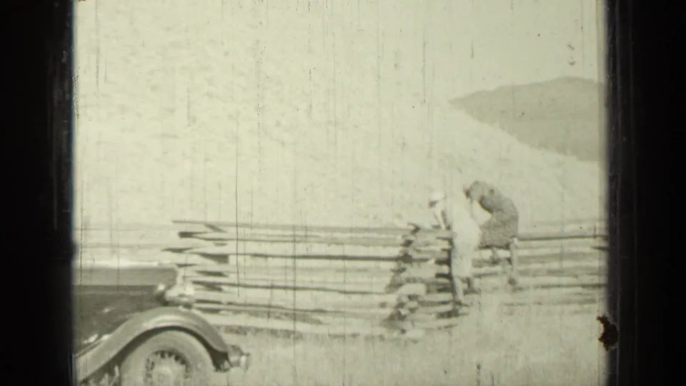 In the Bend, Oregon 1937 scene, two women stand or bend near a rail fence in an open outdoor setting, with part of a car entering the frame.