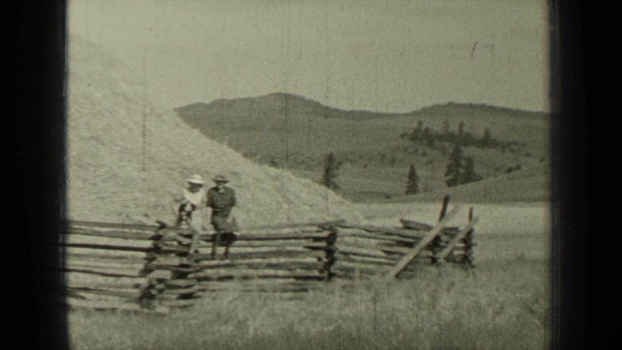Two adults stand beside a split-rail fence in a grassy field with hills behind them.