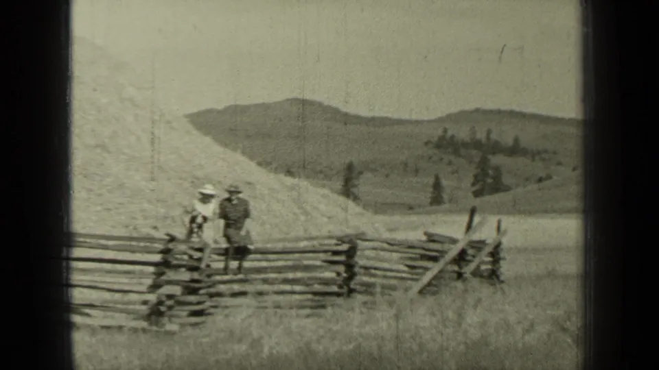 In 1937 Bend, Oregon home-movie footage, two adults in period clothing stand by a split-rail fence in an open grassy field.