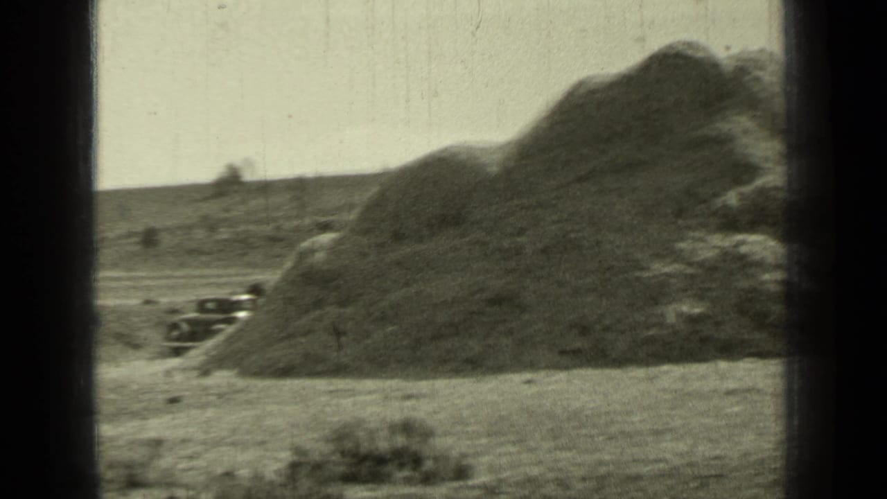 Rural field with a large mound, an older car at left, and dark 8mm frame edges.