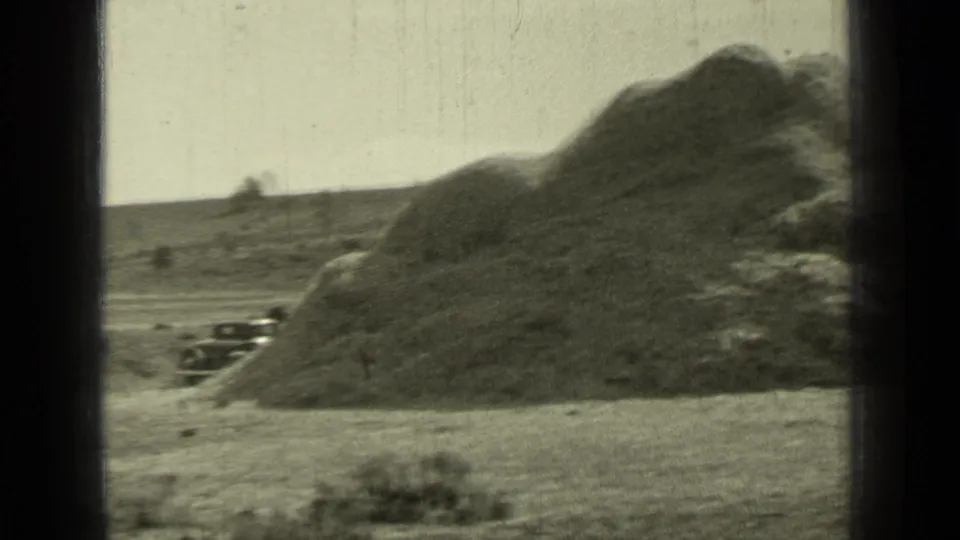 1937 Bend, Oregon home-movie frame showing a large rural mound, an older car partly visible at left, open ground, and distant trees.