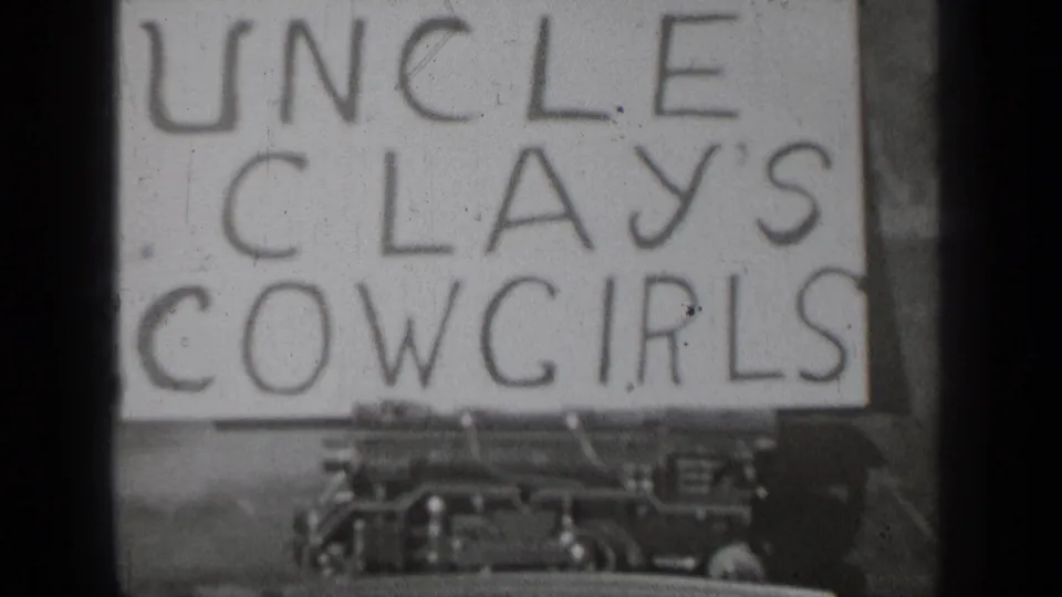 1937 home-movie frame from Bend, Oregon showing a handwritten Uncle Clays Cowgirls sign above the front of a parked automobile.