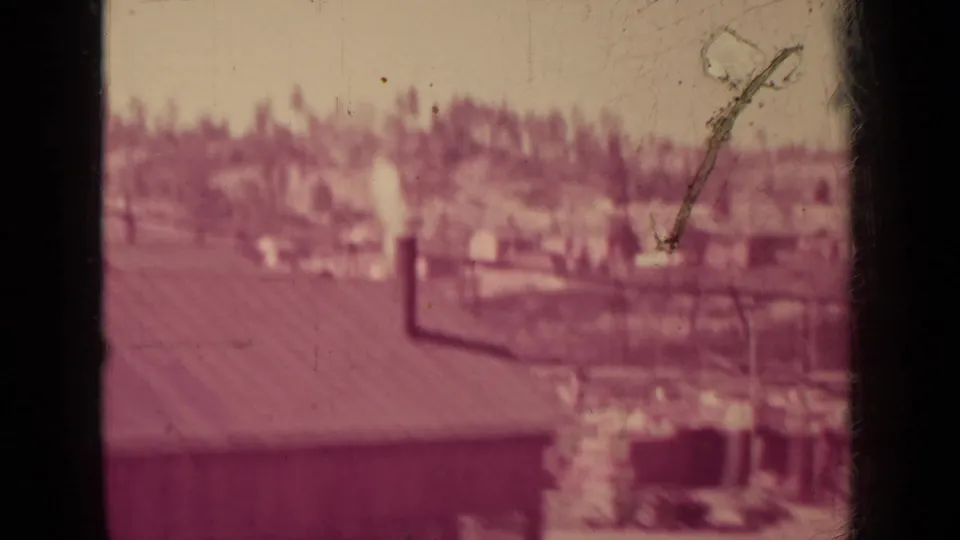 A 1937 Klamath industrial yard appears in soft focus, with low buildings, a smokestack, and trees in the distance.