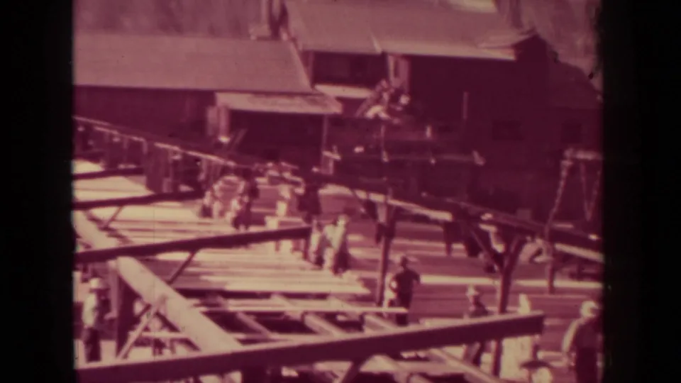 1937 Klamath 8mm frame showing workers in a lumber work yard with elevated rails or conveyors, planks, and industrial buildings.