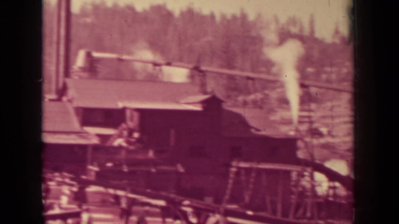 Lumber mill buildings with a smokestack and pale plume near a forested hillside.