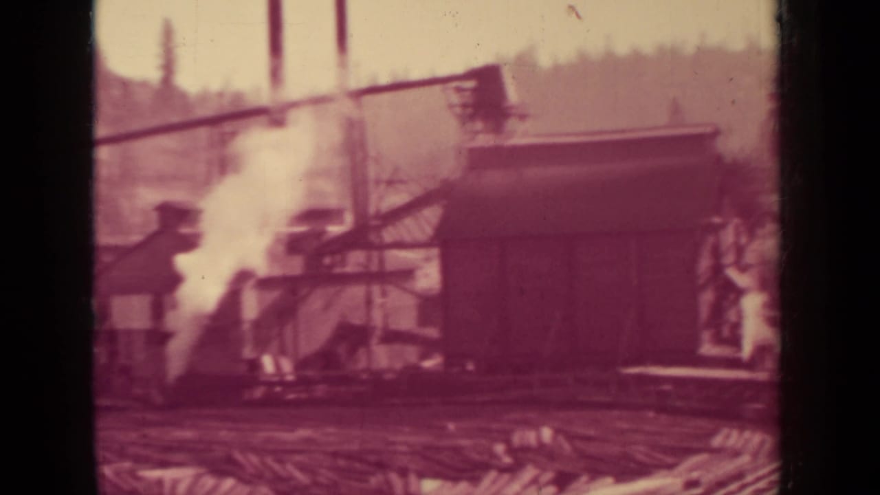 Reddish tinted mill yard with small buildings, steam, overhead cables, and stacked timber.