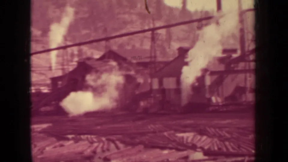 Reddish 8mm frame of the Klamath lumber mill showing steam around low buildings, overhead cables, and timber piles.
